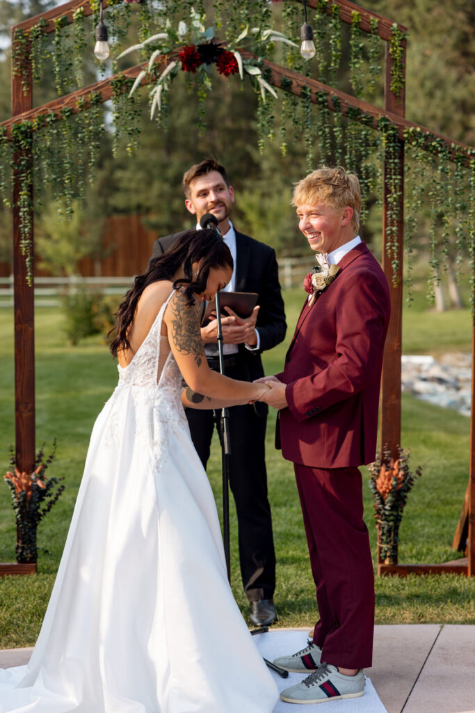 Bride bowing her head laughing while placing the ring on the groom's hand during their outdoor ceremony at Haymoon Resort, officiant smiling behind them — Mikayla + Cayden — Haymoon Resort Wedding — Whitefish Montana — Stacy Edwards Photography