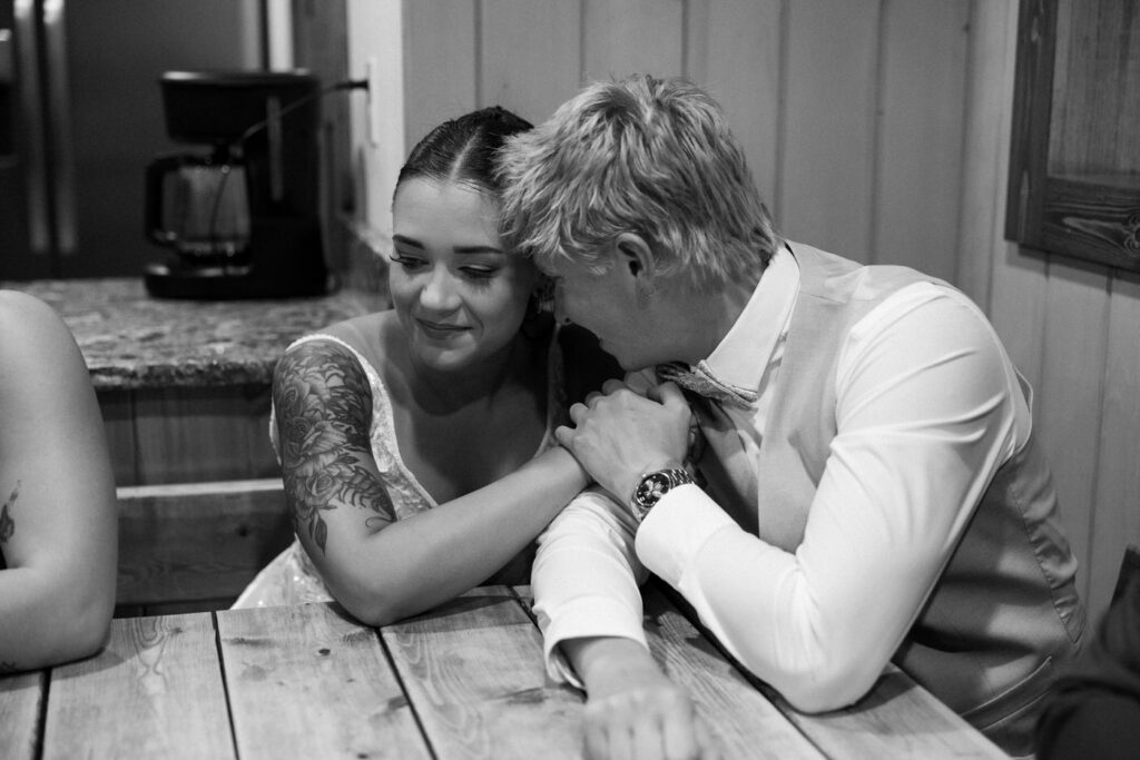 Black and white candid of bride and groom leaning together at a wooden table late in the evening, an intimate quiet moment — Mikayla + Cayden — Haymoon Resort Wedding — Whitefish Montana — Stacy Edwards Photography