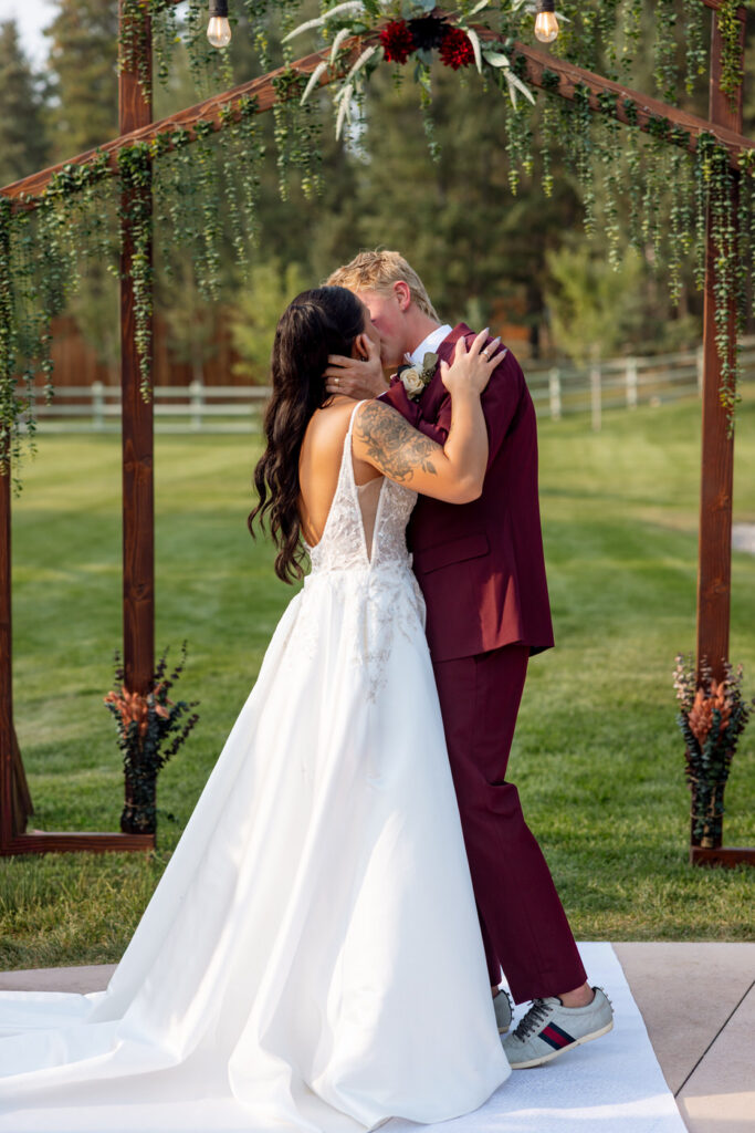Bride and groom sharing their first kiss beneath the wooden greenery arch at Haymoon Resort, her low-back lace gown and floral tattoo visible, white fence beyond — Mikayla + Cayden — Haymoon Resort Wedding — Whitefish Montana — Stacy Edwards Photography