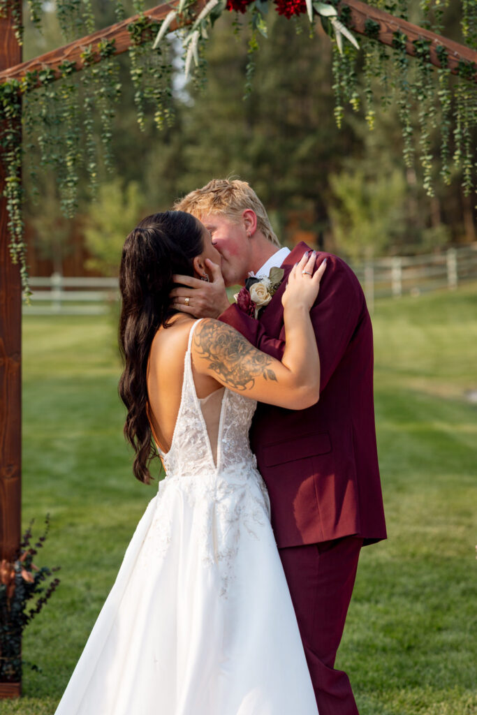 Bride and groom kissing beneath the cascading greenery arch at Haymoon Resort, a closer angle showing her floral arm tattoo and his burgundy boutonnière — Mikayla + Cayden — Haymoon Resort Wedding — Whitefish Montana — Stacy Edwards Photography