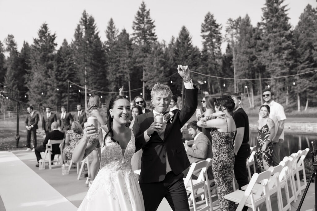 Black and white photo of newlyweds walking back down the aisle holding drinks and pumping a fist in celebration, guests cheering on both sides — Mikayla + Cayden — Haymoon Resort Wedding — Whitefish Montana — Stacy Edwards Photography