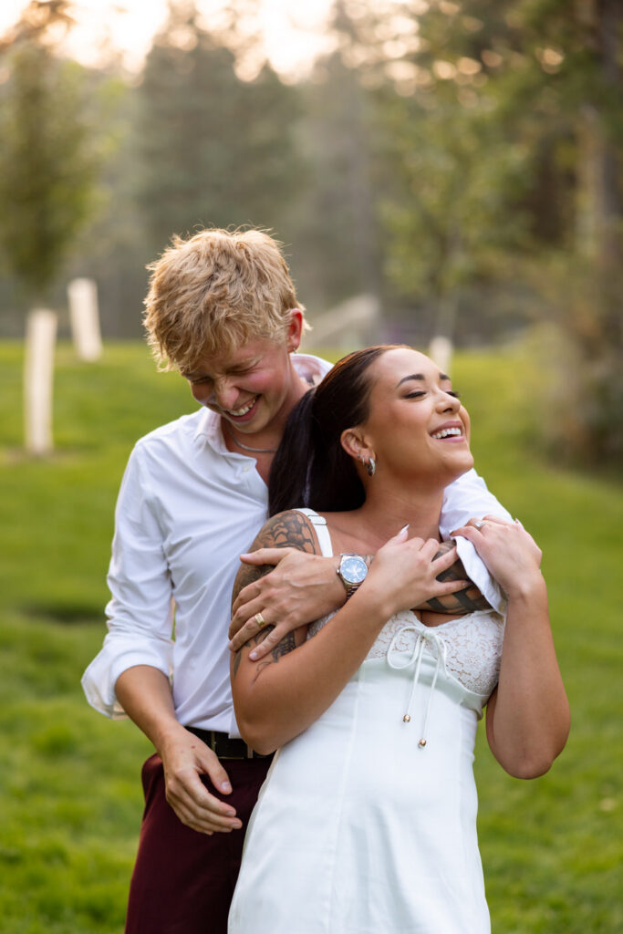 Groom hugging bride from behind as they both laugh on the Haymoon Resort lawn during reception portraits, she in her white mini reception dress and cowboy boots — Mikayla + Cayden — Haymoon Resort Wedding — Whitefish Montana — Stacy Edwards Photography