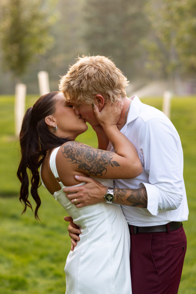 Bride and groom kissing on the Haymoon Resort lawn during golden hour portraits, she in her white mini reception dress and cowboy boots, his hand around her waist — Mikayla + Cayden — Haymoon Resort Wedding — Whitefish Montana — Stacy Edwards Photography