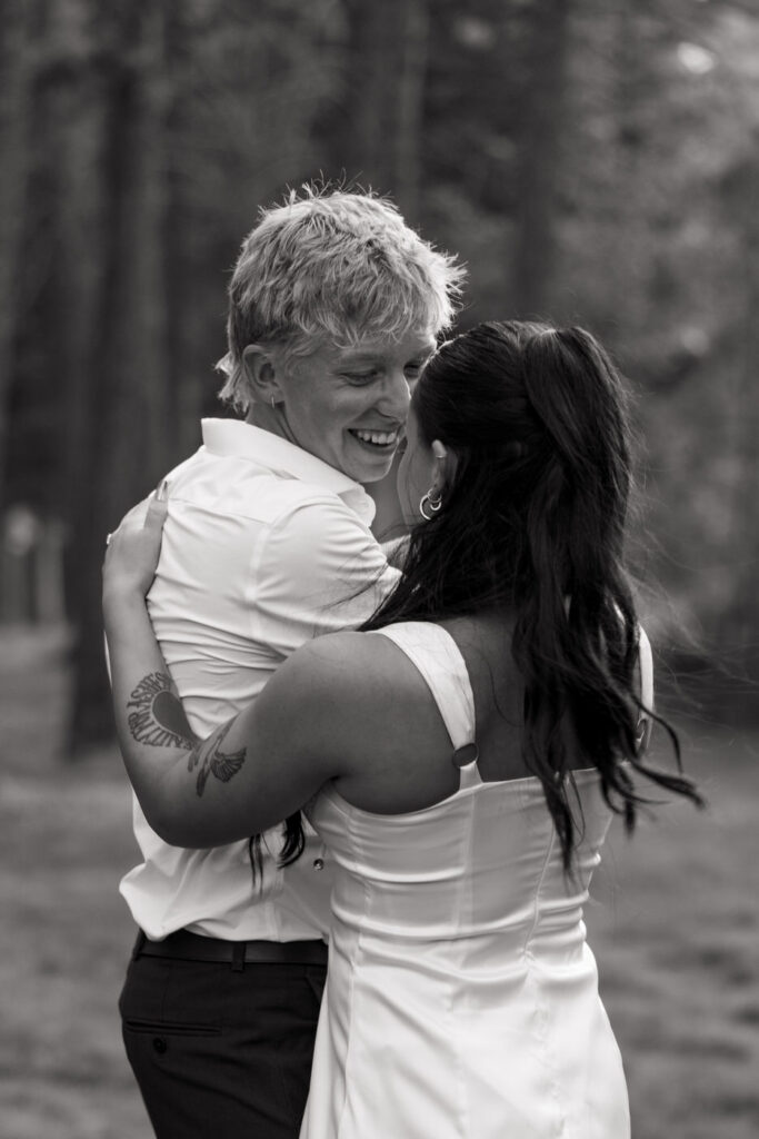 Black and white portrait of groom laughing as bride faces him with arms wrapped around him in the pine trees at Haymoon Resort — Mikayla + Cayden — Haymoon Resort Wedding — Whitefish Montana — Stacy Edwards Photography