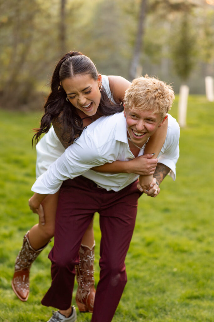 Groom giving bride a piggyback ride on the green grass, both laughing openly, she in cowboy boots and reception outfit — Mikayla + Cayden — Haymoon Resort Wedding — Whitefish Montana — Stacy Edwards Photography