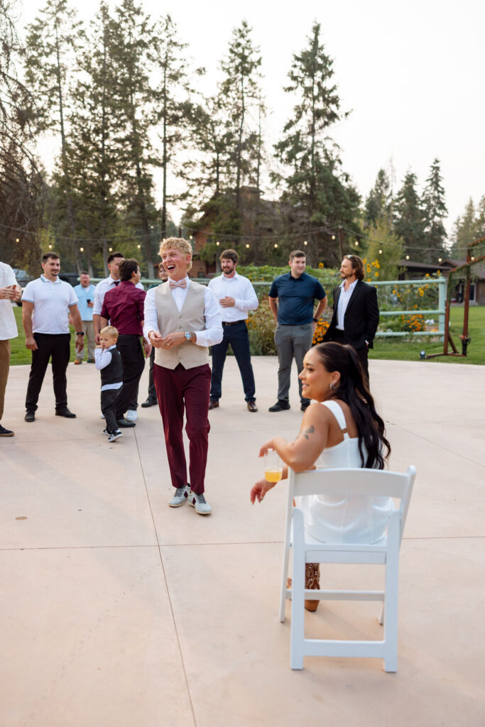 Groom laughing and approaching the bride as she sits in a chair during the garter toss tradition on the outdoor Haymoon Resort dance floor, male guests gathered behind him — Mikayla + Cayden — Haymoon Resort Wedding — Whitefish Montana — Stacy Edwards Photography