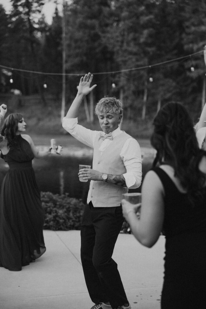 Black and white candid of groom dancing freely on the outdoor Haymoon Resort dance floor with one hand raised, guests and the pond visible behind him — Mikayla + Cayden — Haymoon Resort Wedding — Whitefish Montana — Stacy Edwards Photography