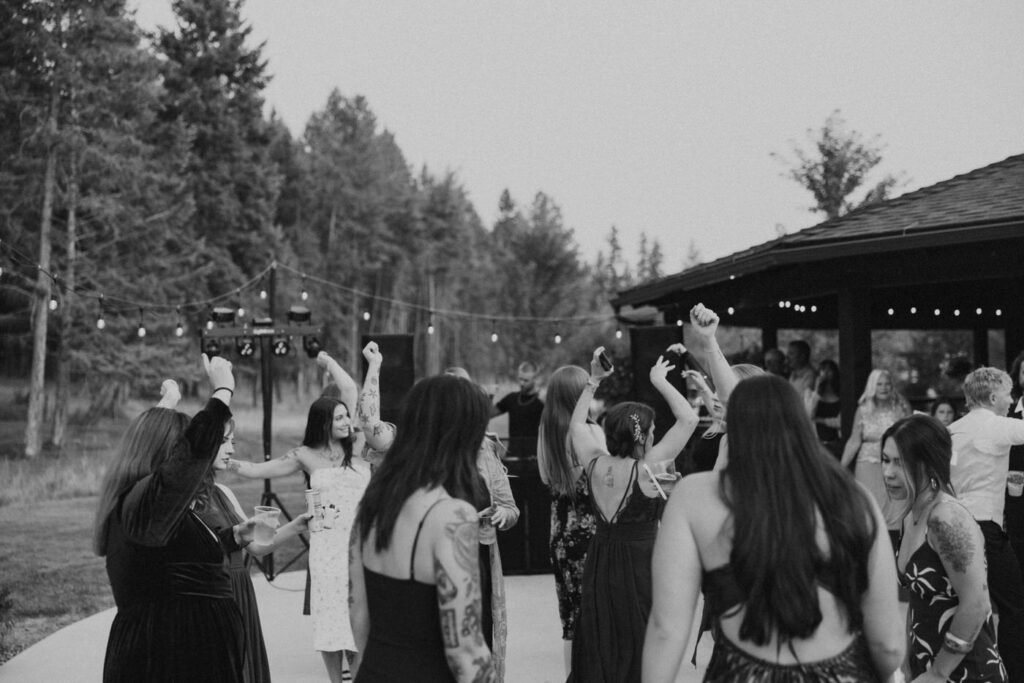 Black and white wide shot of wedding guests crowding the outdoor Haymoon Resort dance floor at dusk, arms raised, DJ setup and Montana pine trees in the background — Mikayla + Cayden — Haymoon Resort Wedding — Whitefish Montana — Stacy Edwards Photography