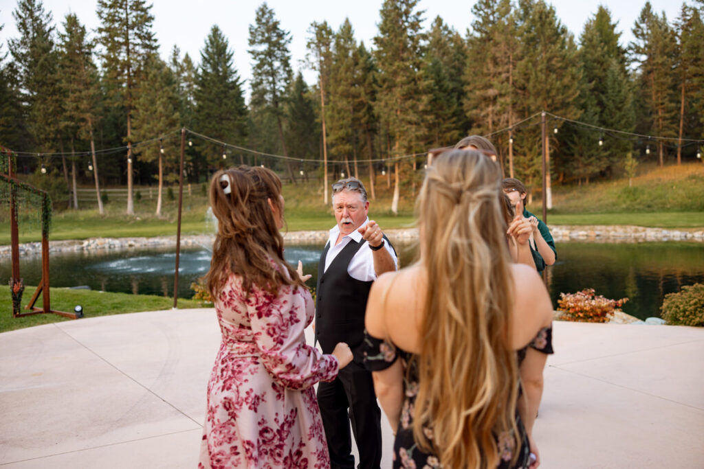 Candid of an older male guest dancing and pointing on the Haymoon Resort outdoor dance floor at dusk, the pond and wooden arch visible behind him — Mikayla + Cayden — Haymoon Resort Wedding — Whitefish Montana — Stacy Edwards Photography
