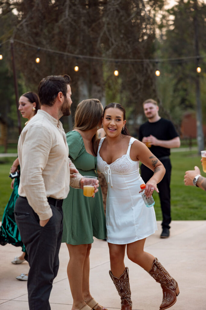 Bride in white reception mini dress and cowboy boots dancing and laughing with guests on the outdoor Haymoon Resort dance floor under string lights at dusk — Mikayla + Cayden — Haymoon Resort Wedding — Whitefish Montana — Stacy Edwards Photography