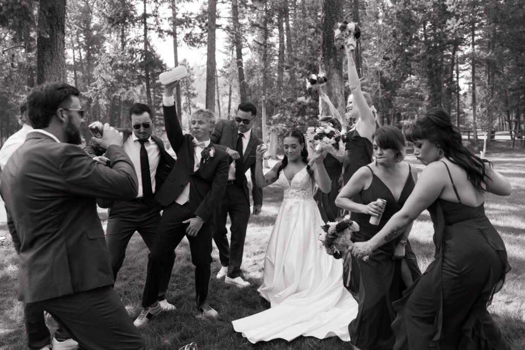 Black and white photo of the full wedding party dancing and celebrating in the Haymoon Resort pine trees, drinks and bouquets raised in the air — Mikayla + Cayden — Haymoon Resort Wedding — Whitefish Montana — Stacy Edwards Photography