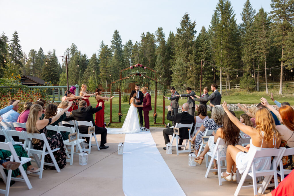 Wide ceremony shot from behind the guests showing bride and groom at the altar beneath the greenery arch, the full wedding party flanking them and bridesmaids reaching their arms toward the couple, guests seated in white chairs on both sides — Mikayla + Cayden — Haymoon Resort Wedding — Whitefish Montana — Stacy Edwards Photography