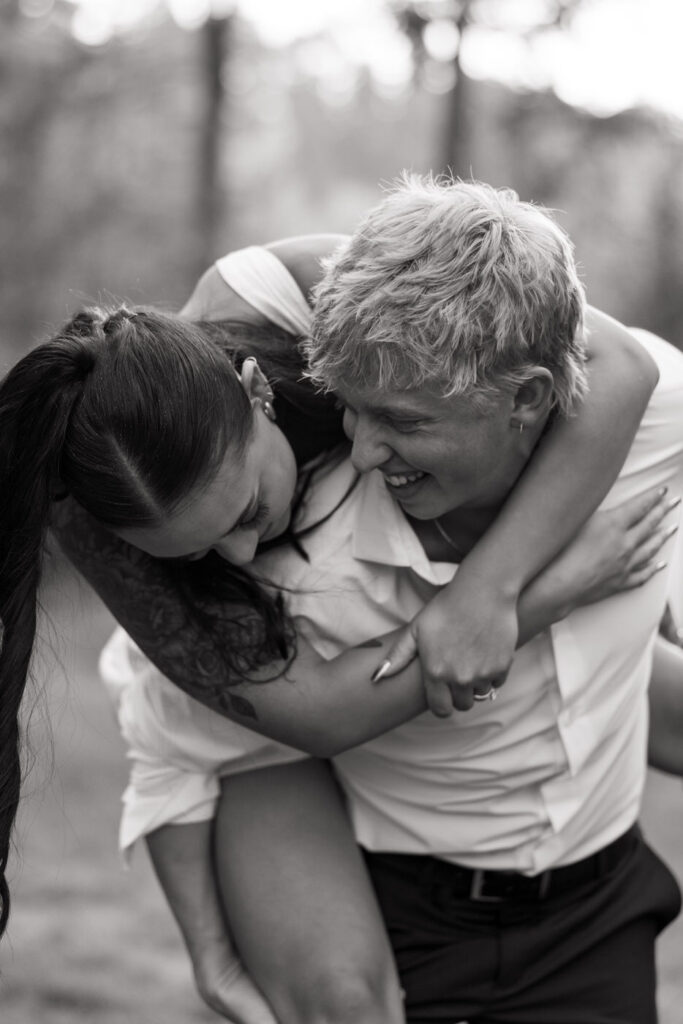 Black and white close photo of bride laughing as groom playfully lifts her in a bear hug in the pine trees at Haymoon Resort, her wedding ring visible on his back — Mikayla + Cayden — Haymoon Resort Wedding — Whitefish Montana — Stacy Edwards Photography