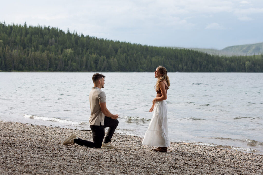 Man on one knee proposing to woman on the rocky shores of Lake McDonald — Ryan + Liz — Surprise Proposal — Glacier National Park — Stacy Edwards Photography