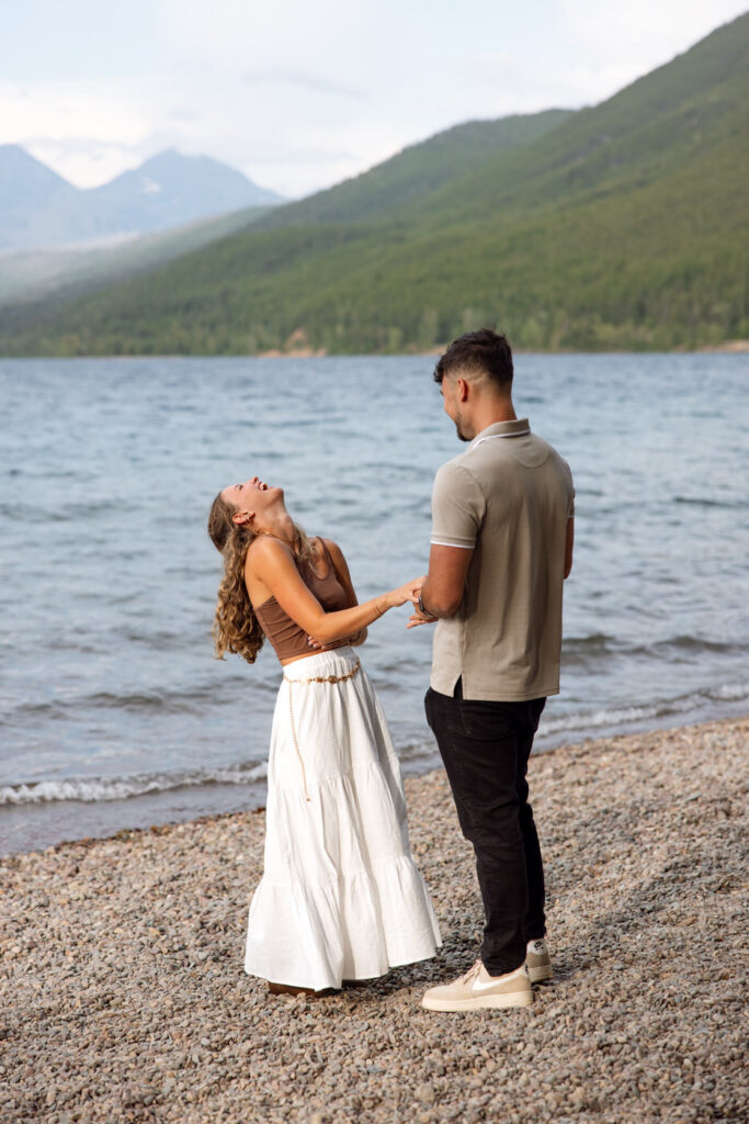 Newly engaged couple laughing together on the shores of Lake McDonald with mountains and blue water in the background — Ryan + Liz — Surprise Proposal — Glacier National Park — Stacy Edwards Photography