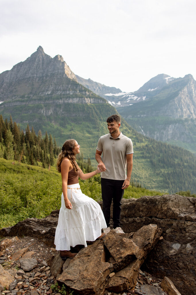 Engaged couple holding hands on rocky overlook along Going to the Sun Road with dramatic Glacier National Park peaks behind them — Ryan + Liz — Surprise Proposal — Glacier National Park — Stacy Edwards Photography
