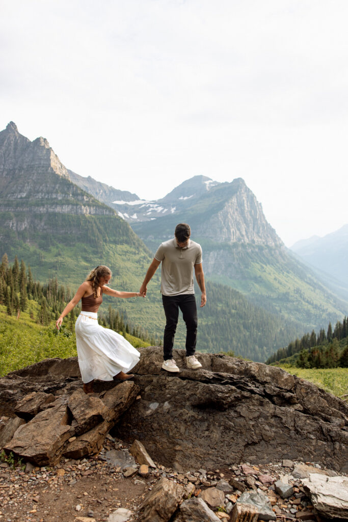 Couple climbing rocks together hand in hand along Going to the Sun Road with sweeping mountain valley views — Ryan + Liz — Surprise Proposal — Glacier National Park — Stacy Edwards Photography