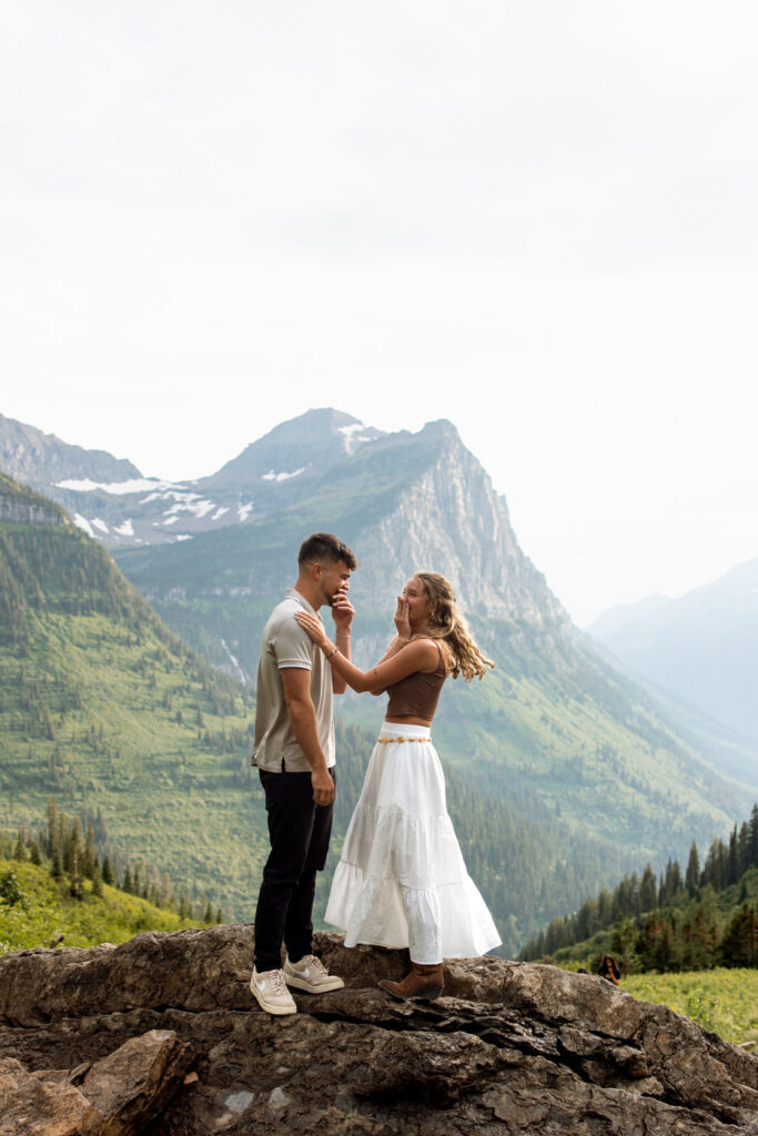 Woman covering her mouth in happy disbelief while couple stands on a rocky overlook with Glacier peaks behind them — Ryan + Liz — Surprise Proposal — Glacier National Park — Stacy Edwards Photography