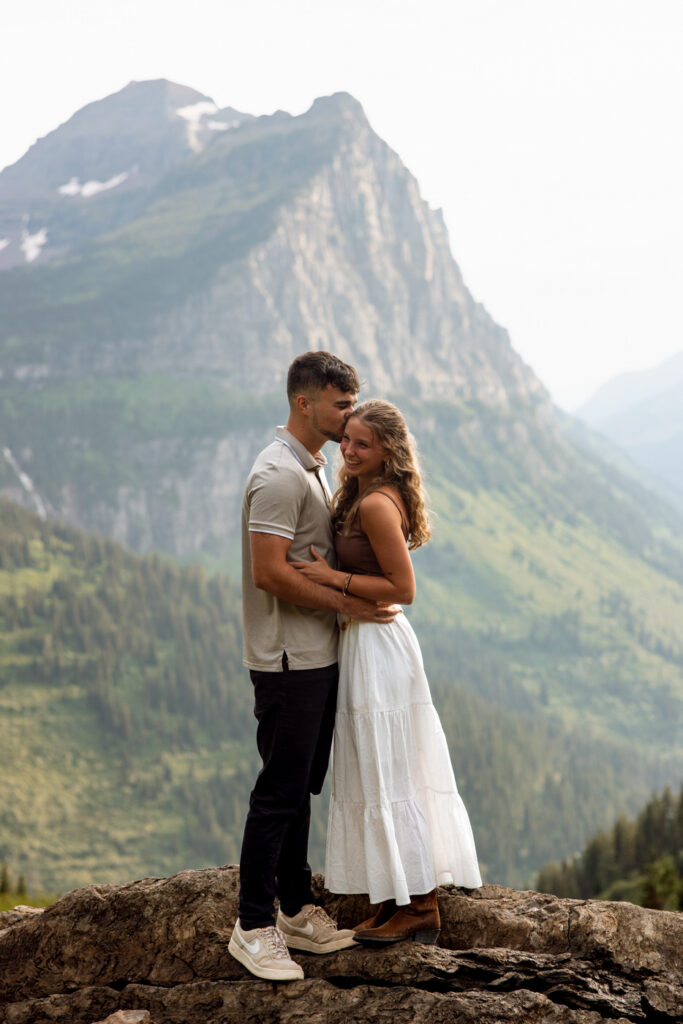 Man kissing woman's temple as she laughs, standing on a rocky overlook with towering Glacier National Park mountains in the background — Ryan + Liz — Surprise Proposal — Glacier National Park — Stacy Edwards Photography