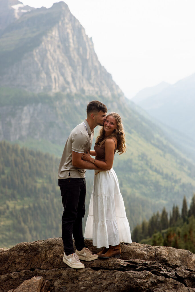Engaged couple cuddling on a rocky overlook at Big Bend, Going to the Sun Road, with misty mountain ridgelines fading into the distance — Ryan + Liz — Surprise Proposal — Glacier National Park — Stacy Edwards Photography