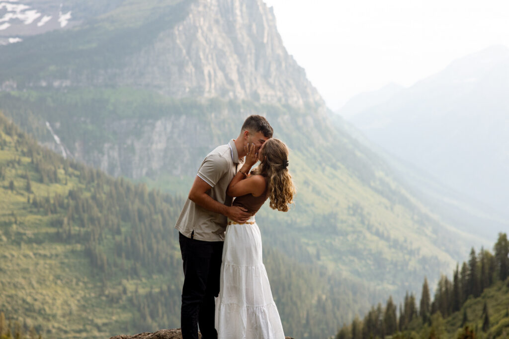 Engaged couple sharing a first kiss on a rocky overlook along Going to the Sun Road with dramatic Glacier National Park peaks and misty valley behind them — Ryan + Liz — Surprise Proposal — Glacier National Park — Stacy Edwards Photography