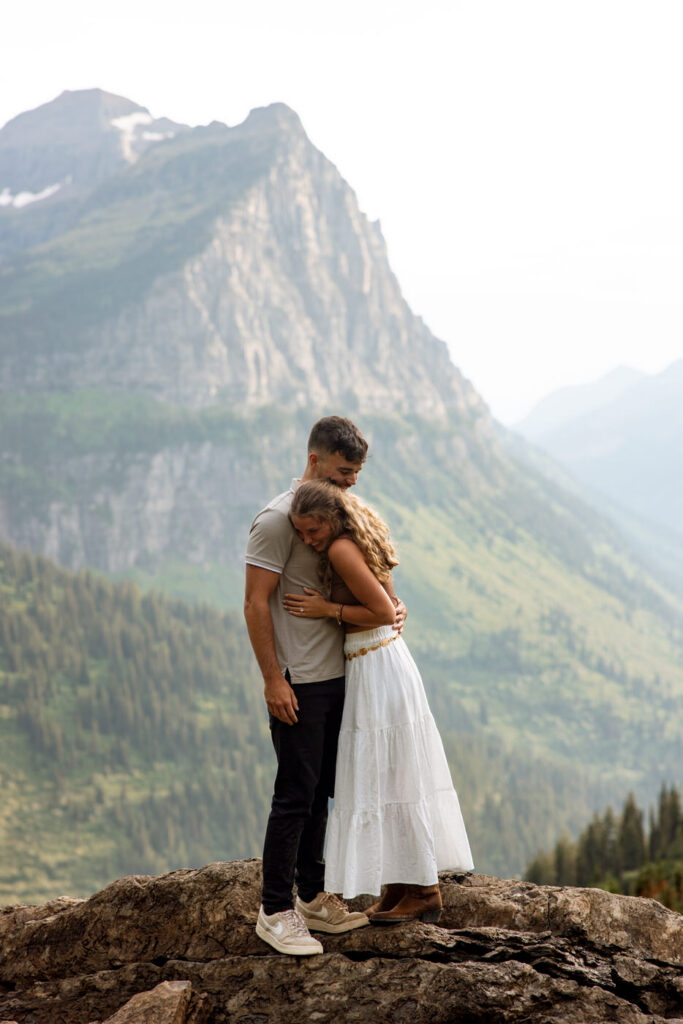 Newly engaged couple sharing a long embrace on a rocky overlook at Big Bend, Going to the Sun Road, with towering Glacier peaks filling the background — Ryan + Liz — Surprise Proposal — Glacier National Park — Stacy Edwards Photography