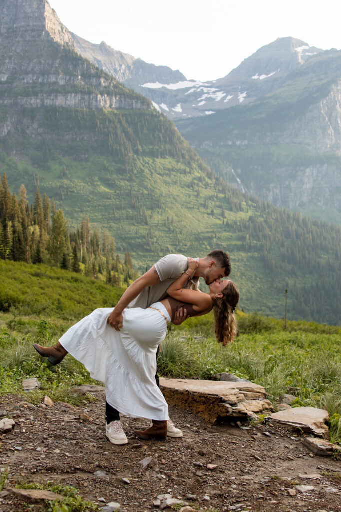 Man dipping woman for a kiss along the Going to the Sun Road trail with sweeping Glacier National Park mountain valley views — Ryan + Liz — Surprise Proposal — Glacier National Park — Stacy Edwards Photography