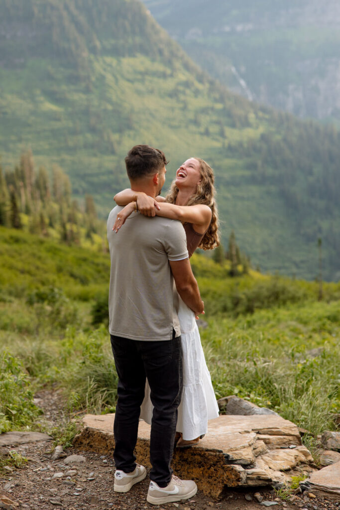 Newly engaged woman laughing with arms around her partner's neck on a hillside overlook along Going to the Sun Road, Glacier National Park — Ryan + Liz — Surprise Proposal — Glacier National Park — Stacy Edwards Photography