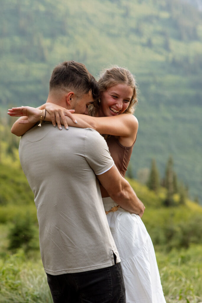 Close-up of newly engaged woman beaming with joy while hugging her partner, lush green Glacier National Park mountainside in the background — Ryan + Liz — Surprise Proposal — Glacier National Park — Stacy Edwards Photography