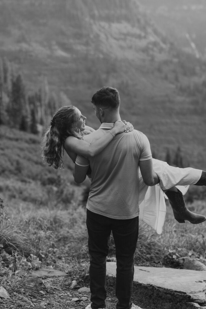 Black and white image of man lifting woman into his arms on a Going to the Sun Road overlook, both laughing, Glacier National Park — Ryan + Liz — Surprise Proposal — Glacier National Park — Stacy Edwards Photography