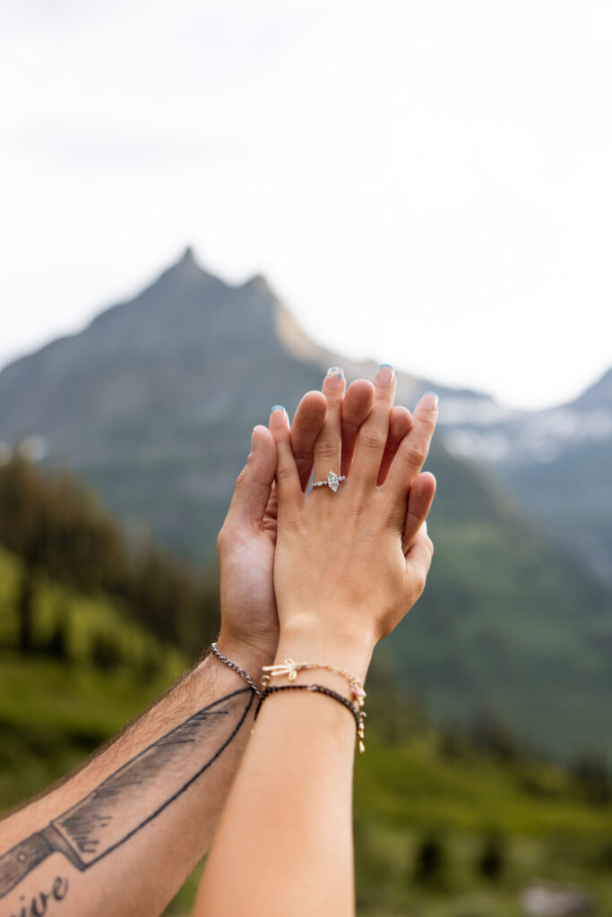 Close-up of engaged couple's hands raised together showing the engagement ring against a Glacier National Park mountain peak backdrop — Ryan + Liz — Surprise Proposal — Glacier National Park — Stacy Edwards Photography