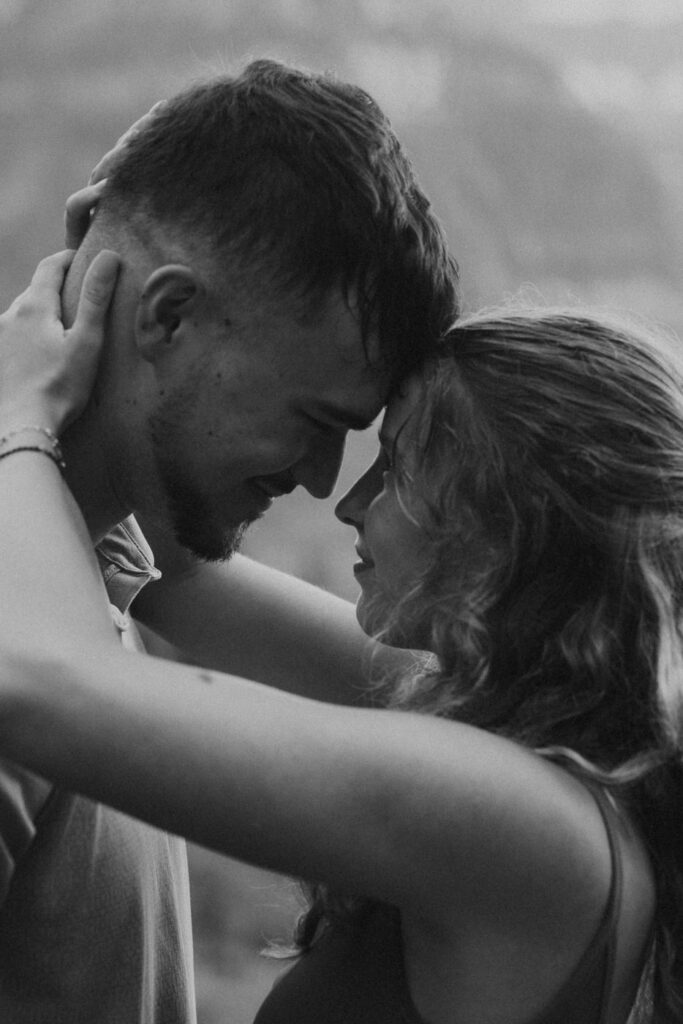 Black and white intimate close-up of newly engaged couple touching foreheads, her hands cradling his face, dramatic cloudy Glacier sky behind them — Ryan + Liz — Surprise Proposal — Glacier National Park — Stacy Edwards Photography