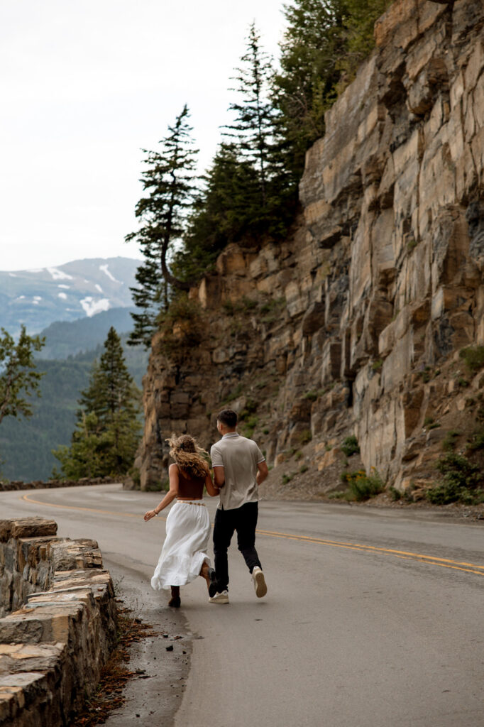 Newly engaged couple running hand in hand along Going to the Sun Road with towering rock cliffs and snow-capped Glacier peaks in the distance — Ryan + Liz — Surprise Proposal — Glacier National Park — Stacy Edwards Photography