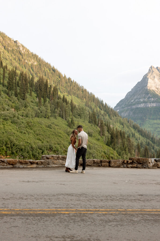 Engaged couple standing close together on Going to the Sun Road at golden hour with forested mountainside and rocky Glacier peak glowing behind them — Ryan + Liz — Surprise Proposal — Glacier National Park — Stacy Edwards Photography