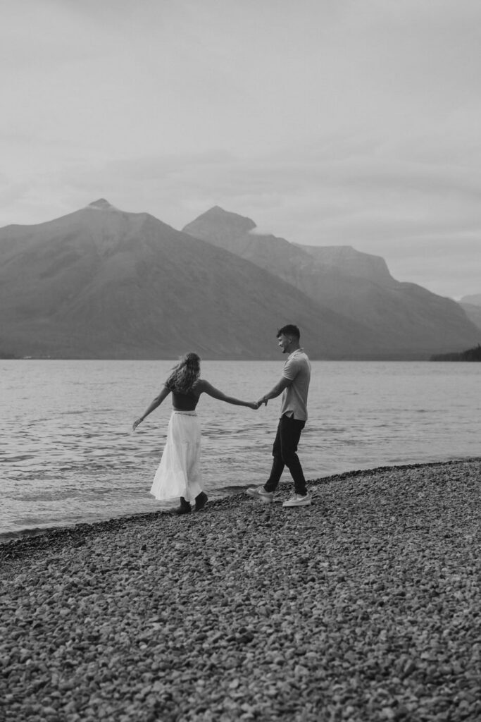 Black and white image of newly engaged couple walking hand in hand along the rocky Lake McDonald shoreline at dusk, mountains reflected in the water behind them — Ryan + Liz — Surprise Proposal — Glacier National Park — Stacy Edwards Photography
