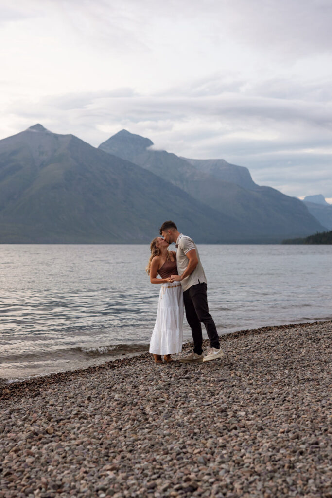 Engaged couple sharing a kiss on the pebbled shores of Lake McDonald at sunset with moody mountain ridgeline in the background — Ryan + Liz — Surprise Proposal — Glacier National Park — Stacy Edwards Photography