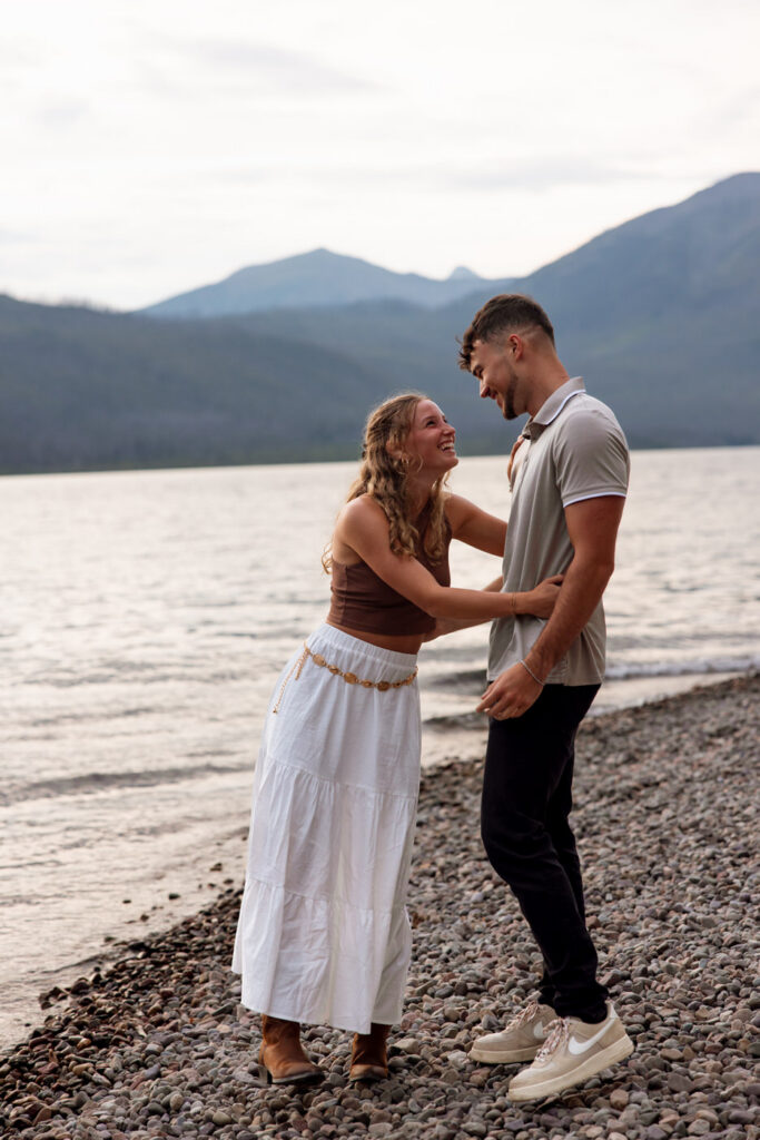 Newly engaged couple laughing and holding hands on the shores of Lake McDonald at golden hour, Glacier National Park mountains behind them — Ryan + Liz — Surprise Proposal — Glacier National Park — Stacy Edwards Photography