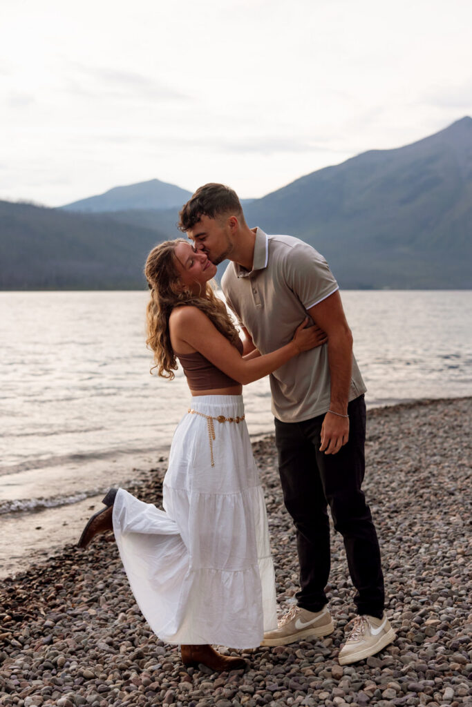 Man kissing woman's cheek as she kicks up her heel on the rocky shoreline of Lake McDonald at dusk, Glacier National Park — Ryan + Liz — Surprise Proposal — Glacier National Park — Stacy Edwards Photography