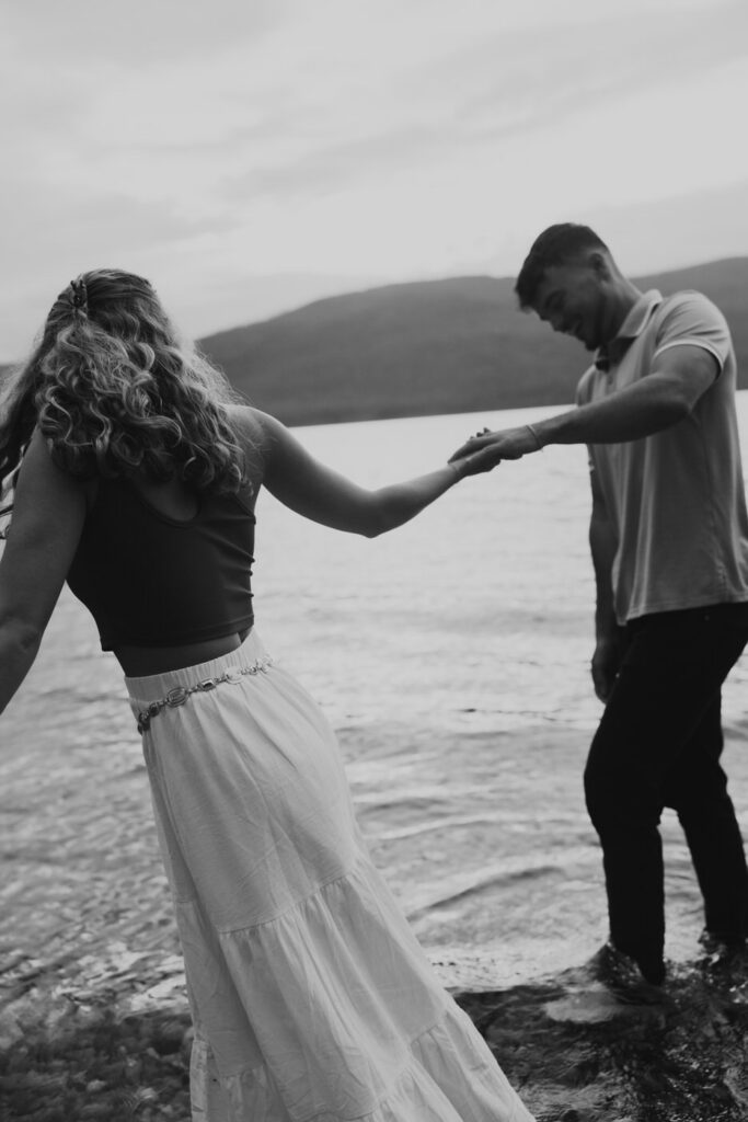 Black and white image of newly engaged couple holding hands at the water's edge of Lake McDonald, mountains visible across the lake — Ryan + Liz — Surprise Proposal — Glacier National Park — Stacy Edwards Photography