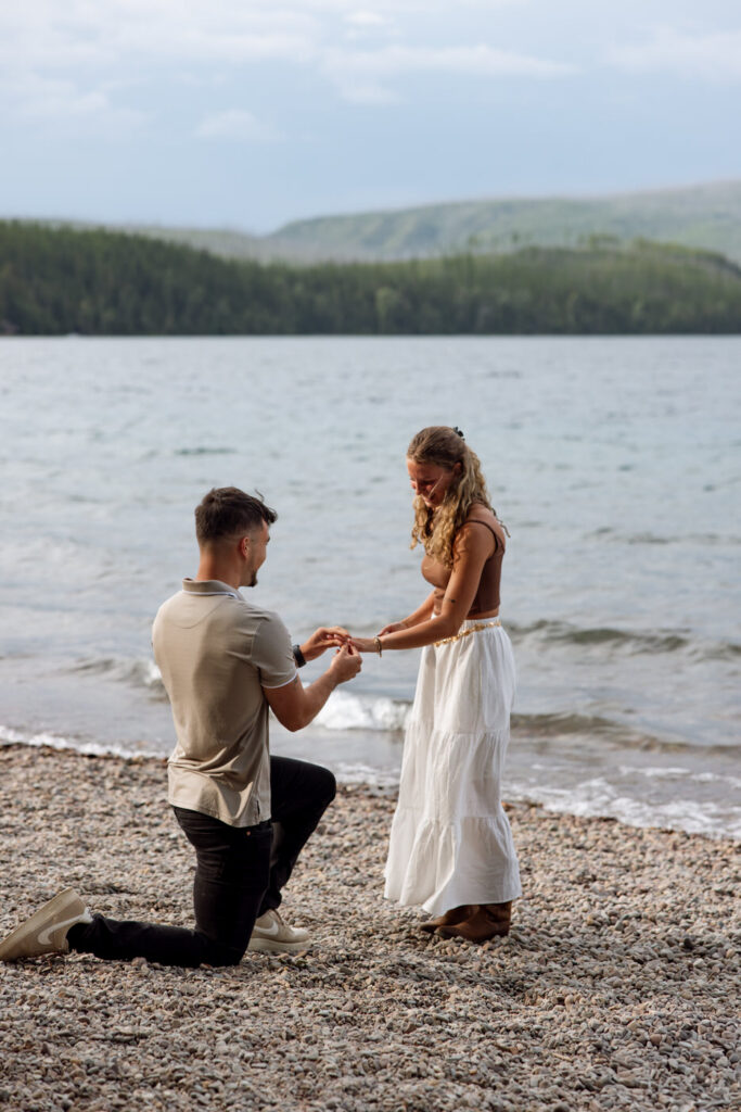 Man placing engagement ring on woman's finger during a lakeside proposal at Apgar Village — Ryan + Liz — Surprise Proposal — Glacier National Park — Stacy Edwards Photography