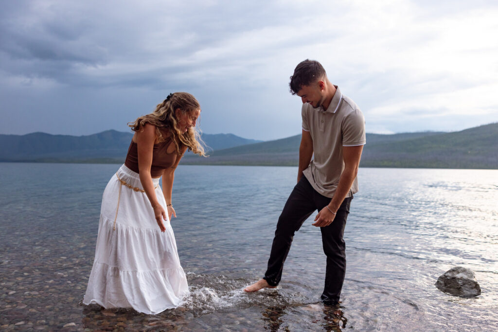 Newly engaged couple laughing and splashing barefoot in Lake McDonald as storm clouds roll in over the mountains behind them — Ryan + Liz — Surprise Proposal — Glacier National Park — Stacy Edwards Photography