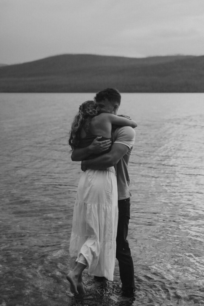 Black and white image of engaged couple embracing and kissing while standing barefoot in Lake McDonald at dusk, soft mountain ridgeline reflected in the calm water — Ryan + Liz — Surprise Proposal — Glacier National Park — Stacy Edwards Photography