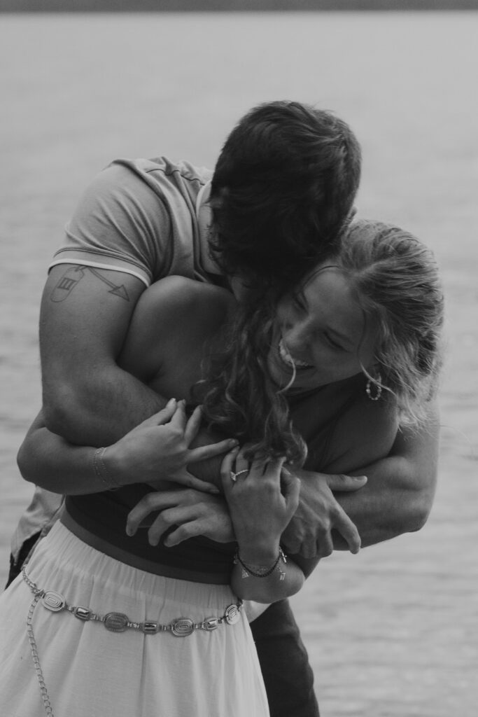Black and white close-up of engaged couple holding each other tightly at the water's edge of Lake McDonald, her engagement ring visible as they embrace — Ryan + Liz — Surprise Proposal — Glacier National Park — Stacy Edwards Photography