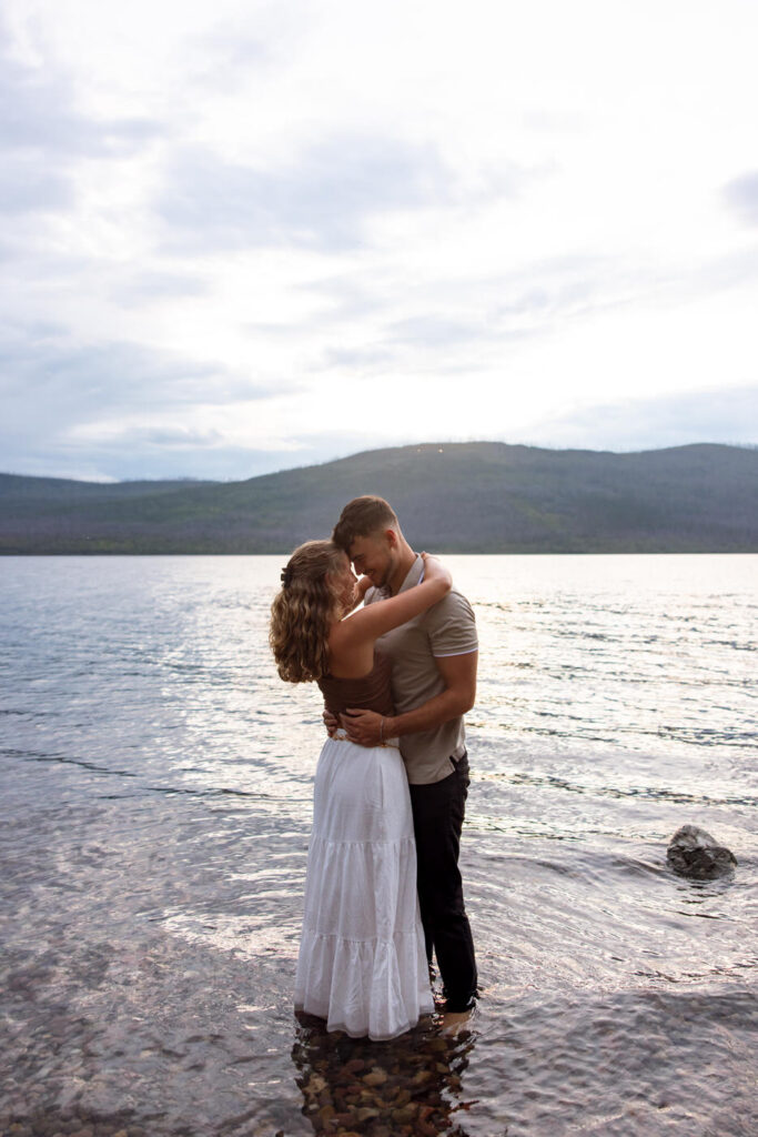 Newly engaged couple touching foreheads and embracing while standing in the glassy shallows of Lake McDonald at sunset, Glacier National Park mountains behind them — Ryan + Liz — Surprise Proposal — Glacier National Park — Stacy Edwards Photography