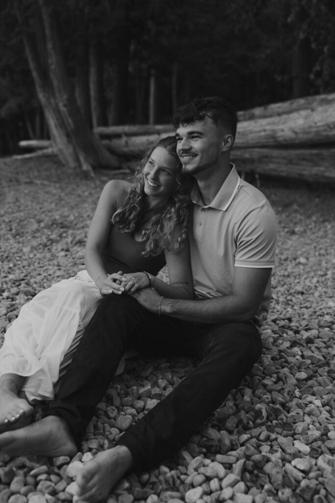 Black and white image of newly engaged couple sitting barefoot together on the pebbled shores of Lake McDonald, laughing and leaning into each other with driftwood logs in the background — Ryan + Liz — Surprise Proposal — Glacier National Park — Stacy Edwards Photography