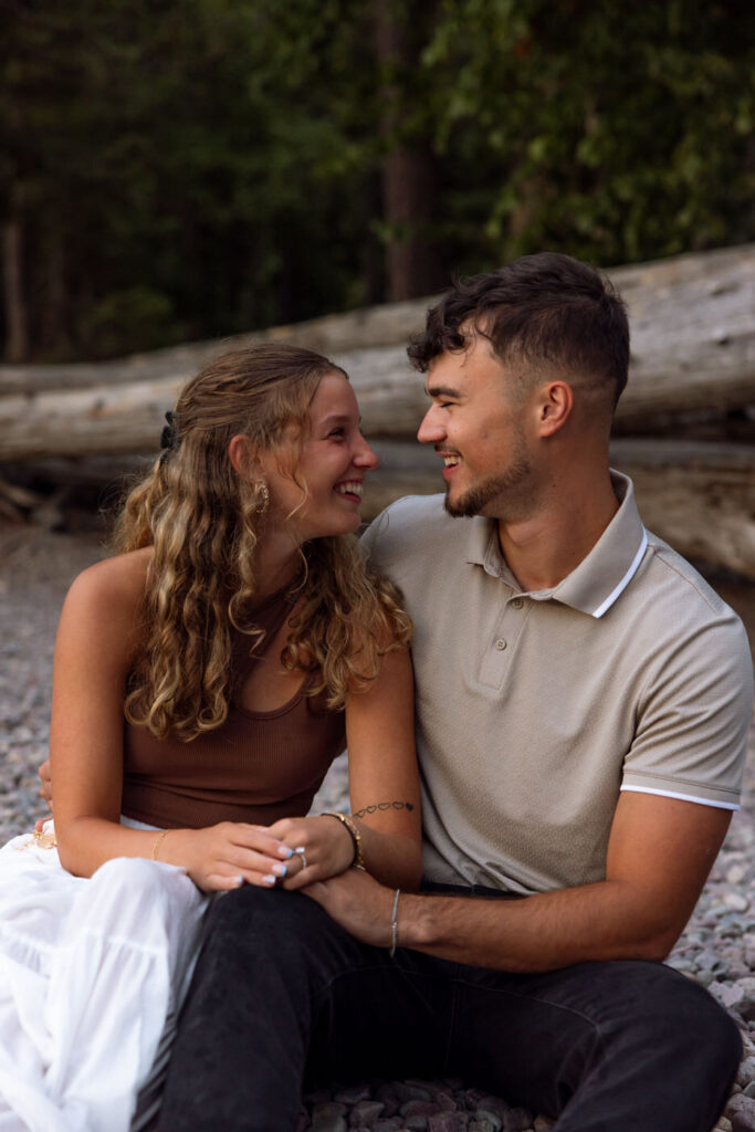 Engaged couple sitting together on the rocky Lake McDonald shoreline laughing and smiling at each other, tall cedar trees in the background, Glacier National Park — Ryan + Liz — Surprise Proposal — Glacier National Park — Stacy Edwards Photography