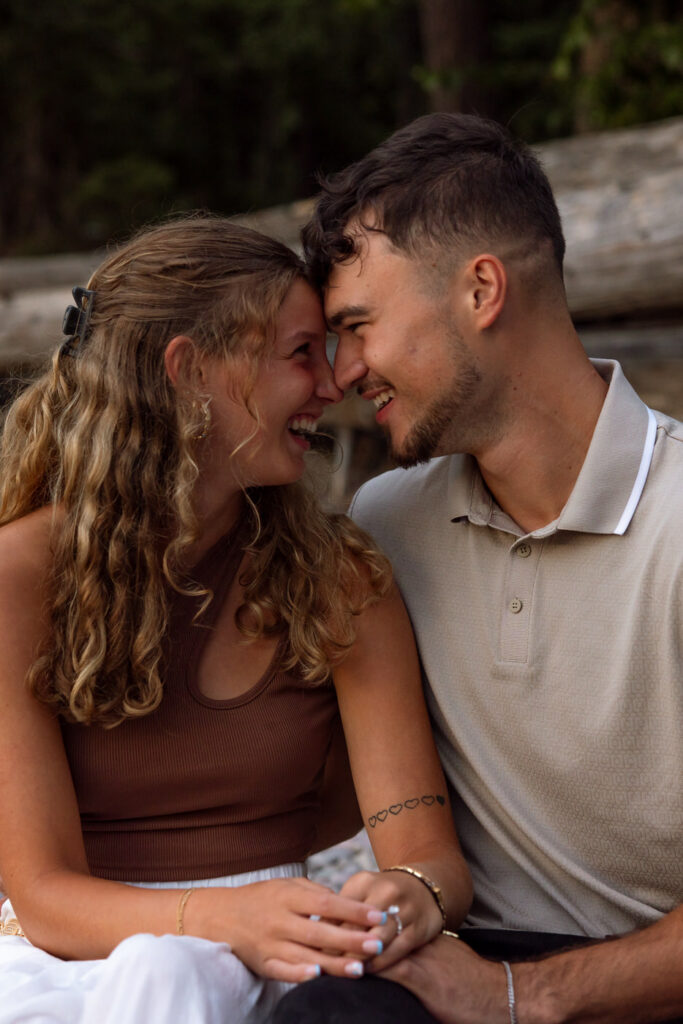 Close-up of newly engaged couple touching foreheads and laughing together while seated on the pebbled shores of Lake McDonald, Apgar Village, Glacier National Park — Ryan + Liz — Surprise Proposal — Glacier National Park — Stacy Edwards Photography