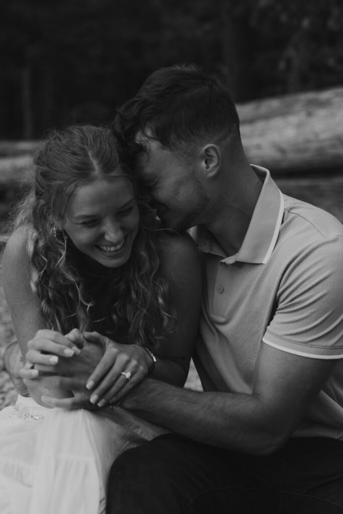 Black and white close-up of man whispering to laughing woman while seated on the rocky shoreline of Lake McDonald, engagement ring visible on her hand — Ryan + Liz — Surprise Proposal — Glacier National Park — Stacy Edwards Photography