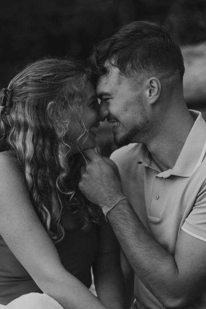 Black and white close-up of newly engaged couple nearly kissing while seated together on the Lake McDonald shoreline at Apgar Village, Glacier National Park — Ryan + Liz — Surprise Proposal — Glacier National Park — Stacy Edwards Photography