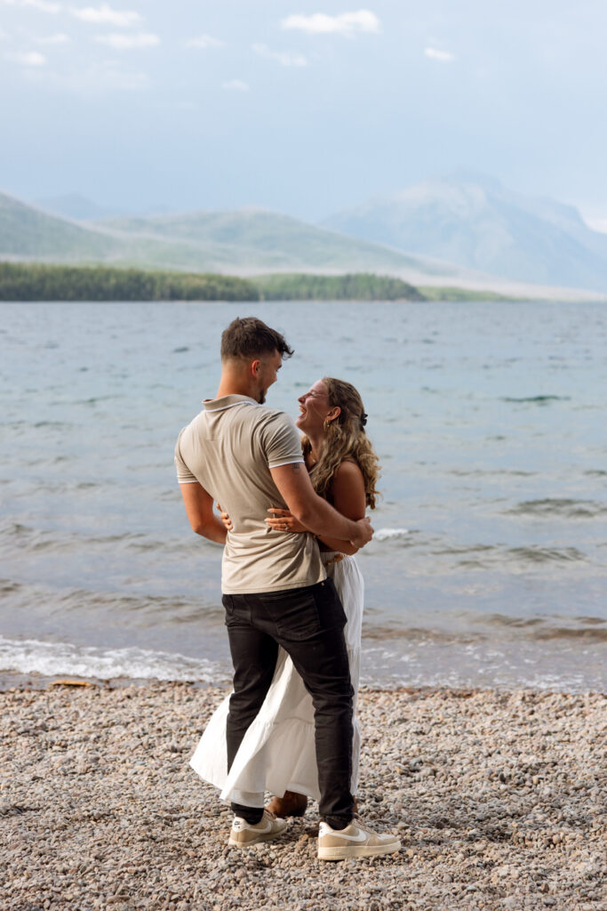 Newly engaged couple laughing and embracing on the shores of Lake McDonald with misty mountains behind them — Ryan + Liz — Surprise Proposal — Glacier National Park — Stacy Edwards Photography